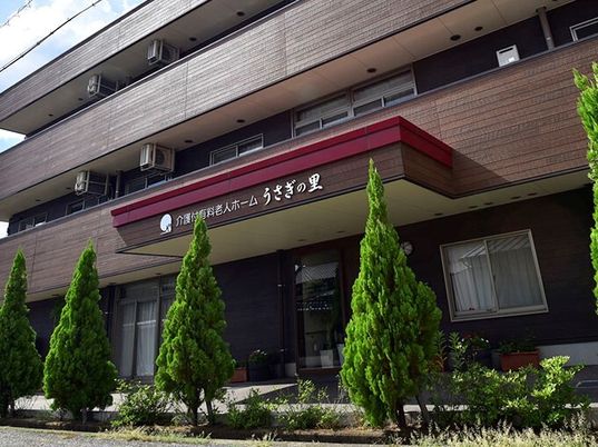 ブラウン系の3階建ての建物は各フロアにベランダがあり、周辺には緑が植えられている。玄関の屋根にはホーム名が書かれている。