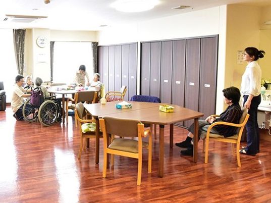 六角形のダイニングテーブルが複数置かれているフローリングの共有スペース。入居者が好きな場所にくつろいでいて、スタッフもいる。