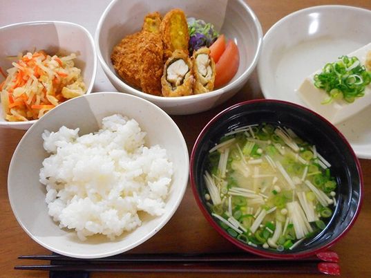 白いご飯に味噌汁、野菜の煮ものに揚げ物、冷ややっこなどが並べられ、手前には箸が置かれている。