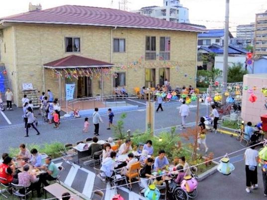 レンガ調の外壁の２階建ての建物にはエンジ系の屋根が付き、玄関には手すりが付いている。お祭りの飾りつけがされ、多くの人でにぎわっている。