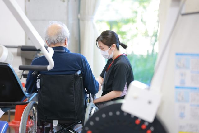 車椅子で訓練を受ける場面 機能訓練室 自然光が入る明るい空間で、車椅子利用者への丁寧なサポートが行われています。
