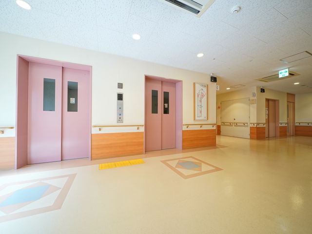 ピンクのエレベーター扉と廊下 エレベーターホール 明るく広々とした空間にピンク色のエレベーターがあり、バリアフリー設計でどなたでも安心してお使いいただけます。