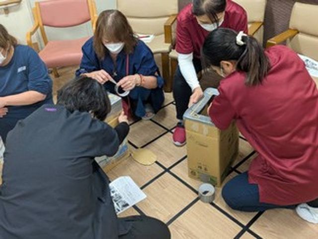 職員が作業する多目的室 多目的室   職員の方々が床に座り、箱を囲んで手作業をされています。準備や整理の様子が見られ、落ち着いて作業しやすい空間です。