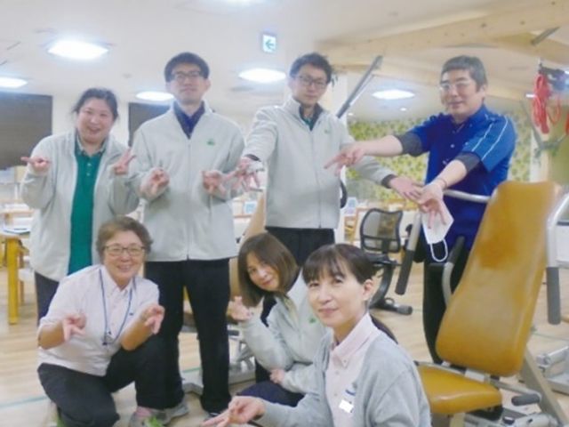 スタッフ集合の機能訓練室 機能訓練室   明るい機能訓練室で、スタッフの方々が笑顔で体操のような動きをしています。働く場の雰囲気が伝わり、チームで支える様子が感じられます。