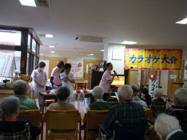 イベントの様子が見える食堂 レクリエーション カラオケ大会が行われており、参加者が楽しんでいる様子が伺えます。職員も一緒になって盛り上がり、和やかな雰囲気が漂っています。