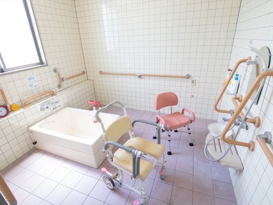 浴室の壁には、濃いベージュ色の手すりが設置されている。ピンク色と黄色のシャワーチェアが置かれている。