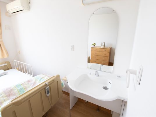 居室にある白い洗面台は、車椅子でのご利用に対応している。鏡は、アーチ形で柔らかい印象を与える。居室にはエアコンも設置されている。