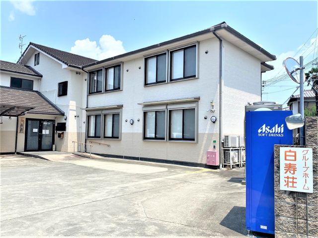 白壁の建物と青い自販機 外観 明るく清潔感のある建物前は、広々としたスペースが確保されており、来訪者のための自動販売機も設置されています。安心して新たな職場を探している方には、心地よい環境が広がっています。