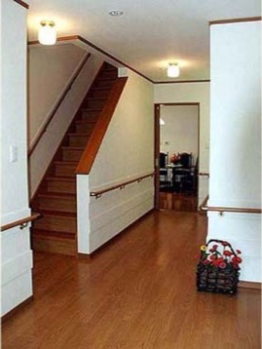 白い壁にフローリングの床である。壁には連続した手すりが付いている。廊下の左手には手すりが付いた階段がある。