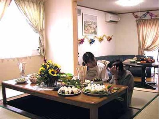 和室の掘りごたつの上には大皿に盛られた料理が置かれている。ミニ向日葵で飾られた花瓶もある。掘りごたつには入居者様とスタッフが座っている。