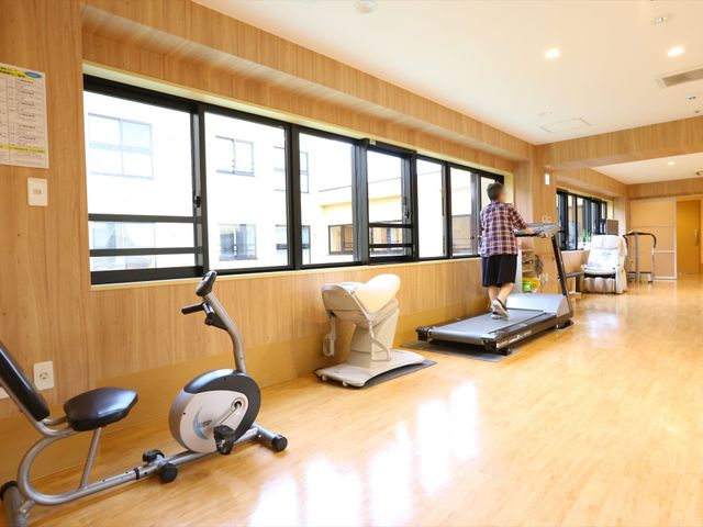 トレーニング機器が並ぶ部屋 フィットネスルーム 明るく広々とした空間で、ご入居者が健康の維持を目指せる設備が整っています。複数のトレーニング機器を使用して、リハビリや運動を行っている様子がうかがえます。