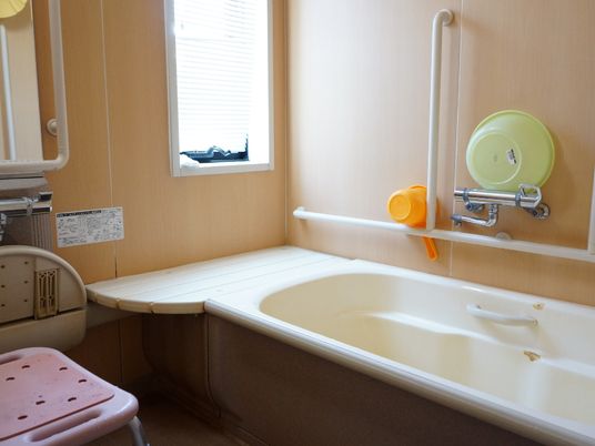 窓から太陽光が差し込む、白とベージュを基調にした明るい浴室。洗い場と浴槽まわりに手すりを設置、シャワーチェアも置かれている。