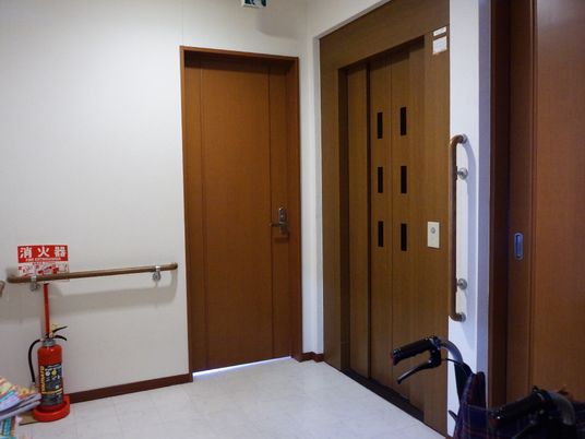 受付の先にエレベーターがある。ドアはブラウンの木目調、左側には非常口、壁には手すりがある。コーナーには消火器が置かれている。