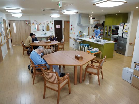 利用者が集っているリビングルームは、大きな照明が備わっていて明るい。非常口を示すサインが天井にある。