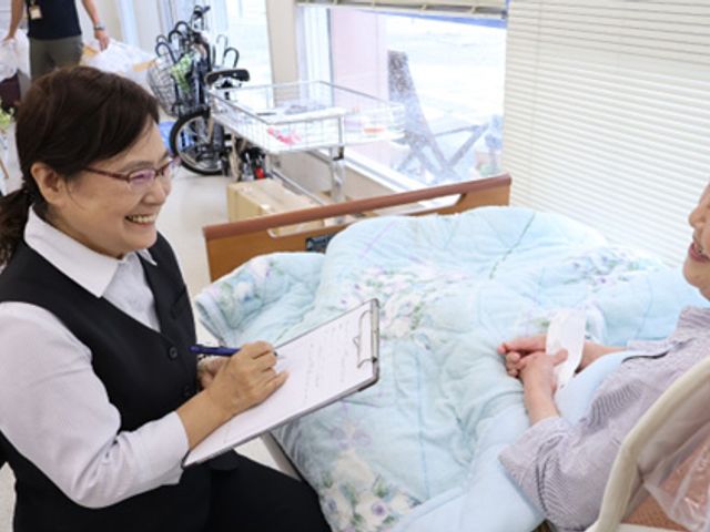 笑顔で話す女性と利用者の居室 居室 明るい居室で、スタッフが利用者と親しげに対話しています。家庭的な雰囲気を大切にしています。