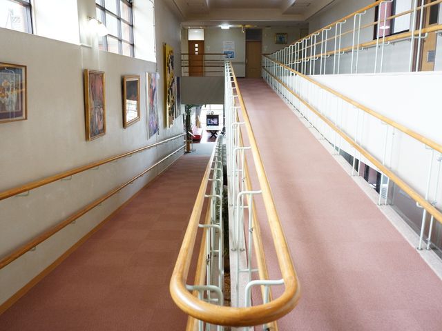 廊下に人がいる様子 室内スロープ ゆったりとした廊下には、壁に絵が飾られ温かい印象を与えています。