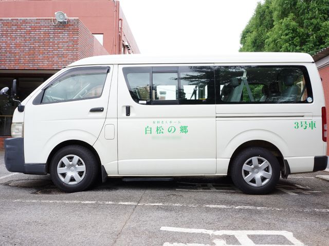 白い送迎車と建物 送迎車 専用の送迎車があり、利用者の移動をサポートしています。安全な乗り降りへ配慮が感じられる車両です。