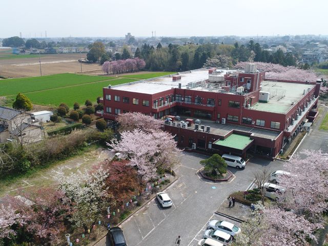 春に咲く桜と施設外観 外観 四季を感じる広々とした景色が自慢の施設です。春先の桜が入居者の憩いのひとときを提供します。