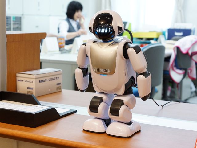 ロボットがカウンター前に立つ 受付窓口 最先端のロボットが受付業務をサポートする姿です。バックで作業するスタッフと共に快適な環境作りに貢献しています。
