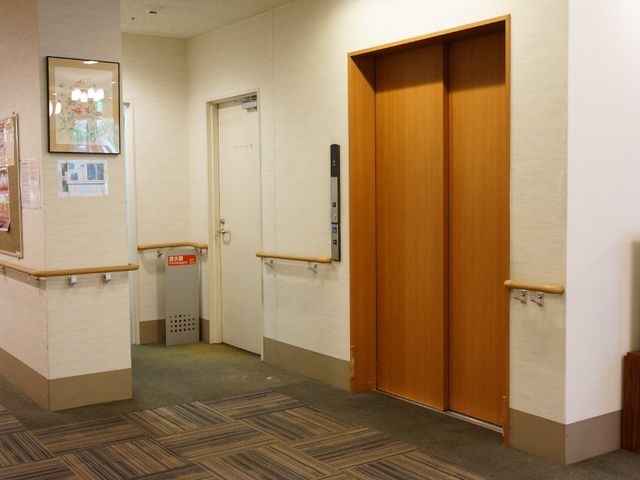 廊下とエレベーター エレベーターホール 廊下はゆったりとした造りで、掲示物がある壁も視覚的に安心感を与えています。エレベーターは手すり付きで、安全に利用できる設計となっています。