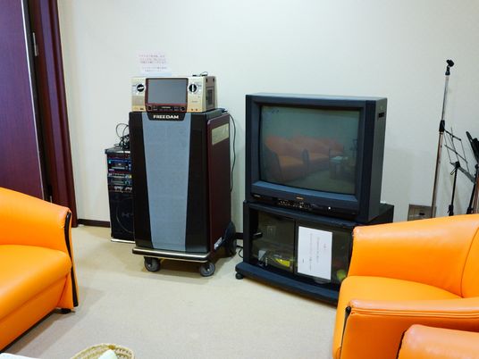 施設の写真 カラオケ室では、部屋にオレンジ色の椅子が用意されている。家庭用のテレビにつなぐカラオケ機器や、マイクスタンドが設置してある。