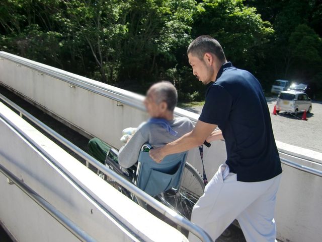 スロープでの介助 外観 日差しの中、車椅子の方をスタッフが丁寧にサポートしています。優しい介助が求められます。