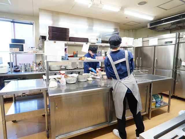 キッチンで働くスタッフ 厨房 清潔感あふれるキッチンで、スタッフが調理に取り組んでいます。私たちと一緒に、健康な食事を支えませんか？