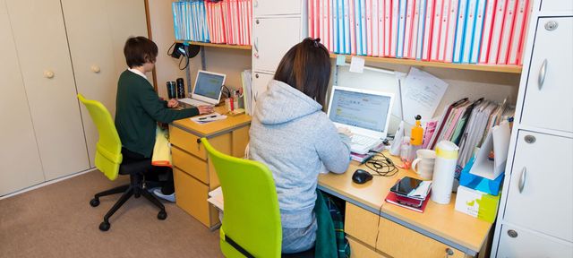 緑の椅子のある居室 居室 ゆとりのある作業スペースで、二人が向かい合わせで快適に過ごしています。明るく整然とした環境が確保されており、集中して作業ができそうです。
