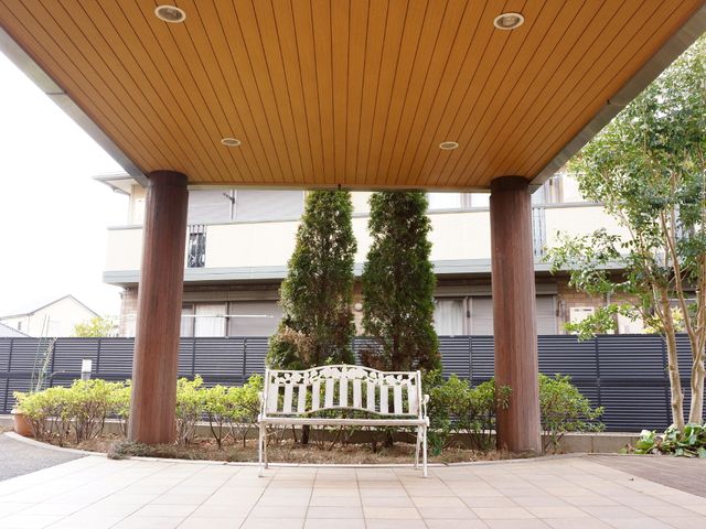 ベンチと樹木のある中庭 エントランス4 屋根付きの休憩スペースで、ゆったりとした時間を過ごせる場所です。白いベンチが設置され、周囲には緑が植えられています。