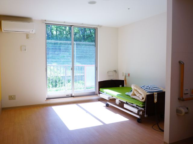 居室のベッドと窓 個室 明るい自然光が差し込む居室です。窓からは緑豊かな景色を望みながら、ゆったりとした時間を過ごすことができます。
