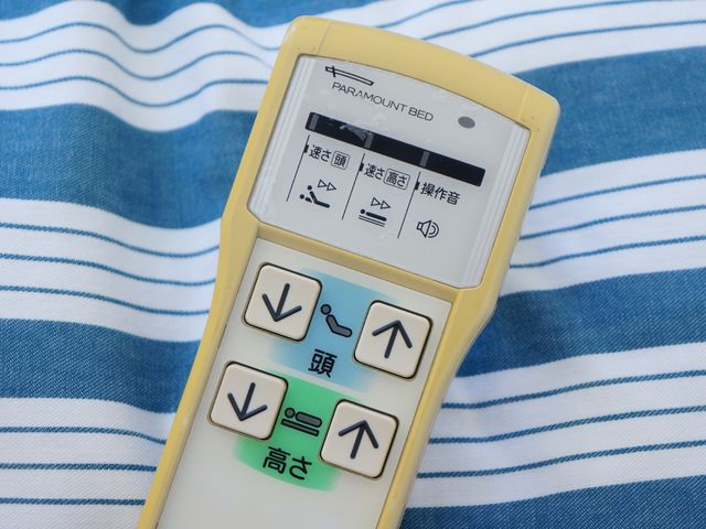 ベッドリモコンとブルーの布 ベッド 各居室にはベッドの高さを調整できるリモコンが完備されており、利用者の快適な生活をサポートしています。
