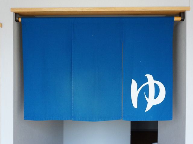 青いのれんが掛かる出入口 大浴場 温かみのあるエントランスに掛けられた青いのれんが、訪れる方を優しくお迎えします。