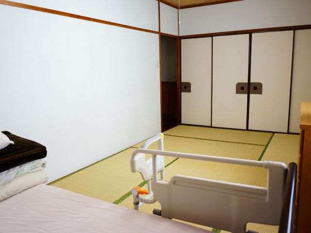 和室の居室風景 和室の様子 落ち着いた雰囲気の和室で、生活の一部としての安らぎを提供しています。手すり付きのベッドが配置され、安心して過ごせる空間となっております。