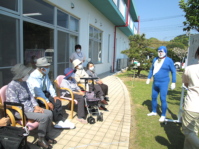 人が集まる庭 庭 庭でのイベント中、利用者が楽しげに座っています。青い着ぐるみのスタッフが話しかけています。