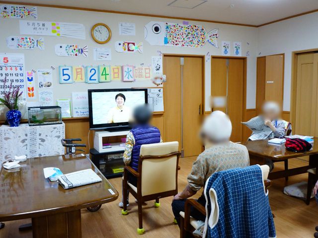 共有スペースでの日常風景 リビング3 壁に飾られたカレンダーや作品が彩りを添える居心地の良い共有リビングです。チェアに座る方々がテレビを視聴しており、和やかな時間が過ごせそうです。
