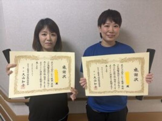 表彰状を持つ職員2人の室内  