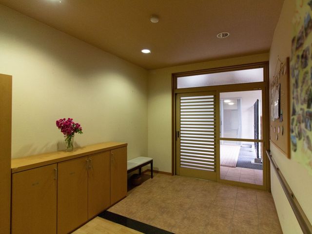 廊下に花とベンチ 玄関 温かみのある色合いで落ち着いた雰囲気のエントランスです。壁には掲示物が飾られ、訪れる人を優しく迎えています。