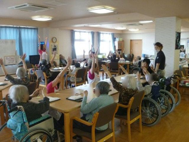 明るい多目的室の様子 レクリエーション 広々とした明るい部屋で、利用者が楽しそうに体操を行っています。スタッフが優しく指導しており、コミュニケーションも活発です。