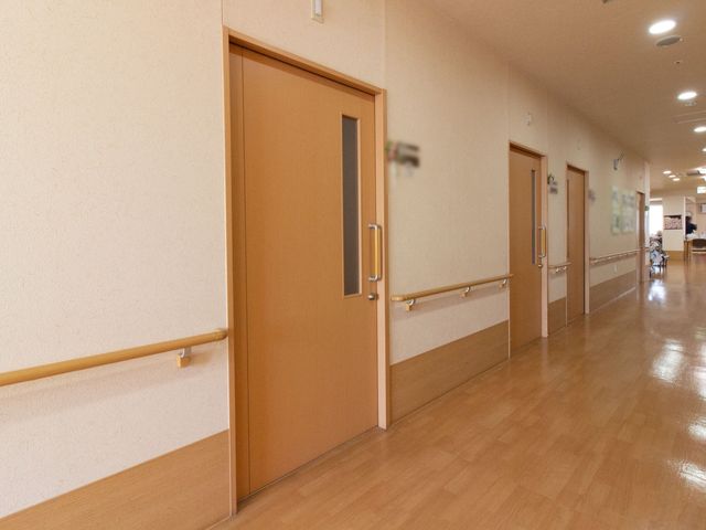 廊下の明るい雰囲気 廊下 ゆとりある廊下が広がり、安全に配慮した手すりが整備されています。遠くにはゆっくりと移動する方の姿も見え、温かな雰囲気が伝わってきます。