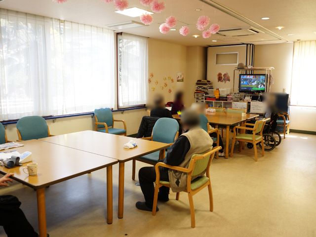 共用スペースでの生活風景 広い居室 ゆったりとしたテーブルと椅子が配置された共用スペースで、利用者の方々がリラックスして過ごされています。明るく開放感のある空間で、デイサービスを提供しています。