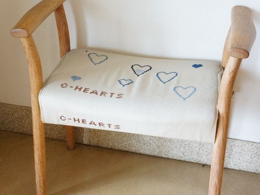 木製の背もたれのない椅子で、座面部分にはクロスステッチの刺繍がされている。施設名とハートなどが刺繍されている。