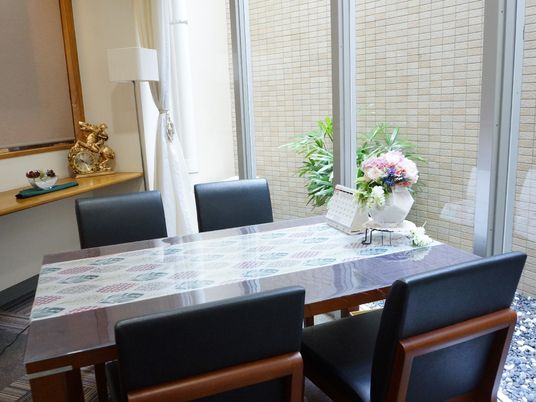 応接室には茶色いテーブルが置かれ、黒い椅子が4つ設置されている。テーブルの上には花瓶に入った花が飾られ、カレンダーも置かれている。