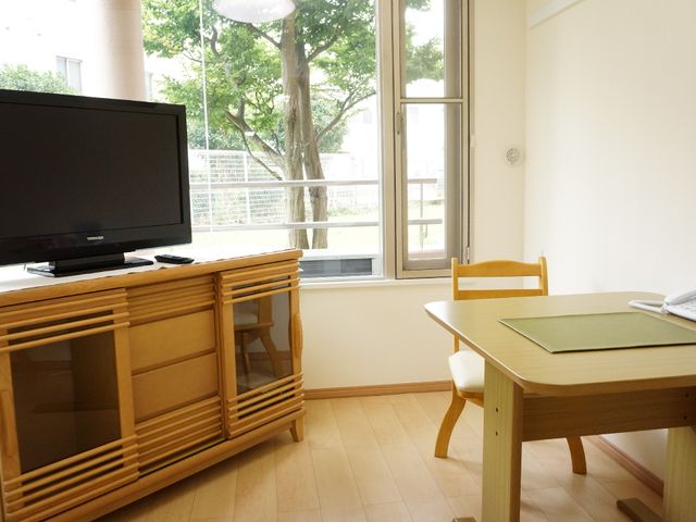 居室内のテレビとテーブル 面会室 落ち着いた雰囲気の居室には、大きなテーブルとテレビが配置され、くつろぎ空間を提供しています。窓からは自然光が入り、心地よい環境です。