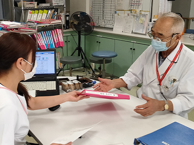 書類を手渡すスタッフ スタッフイメージ 職員が資料をやり取りする様子をご覧いただけます。チームワークを大切にする環境です。