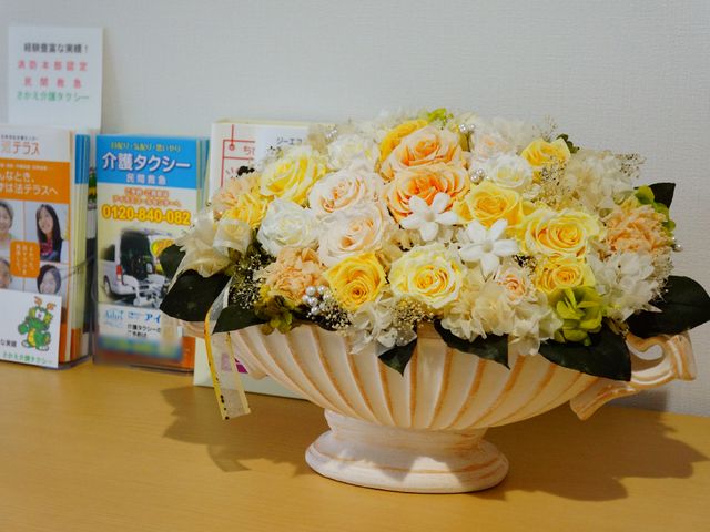 フラワーアレンジメントのディスプレイ 棚の上 温かみのある色合いのフラワーアレンジメントが、暖かな歓迎の気持ちを表しています。バックには施設の案内パンフレットが備えられており、来訪者に情報を提供しています。