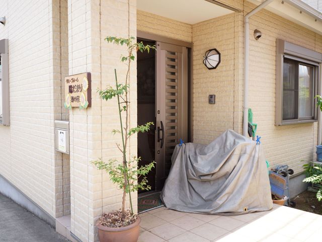 明るいエントランスの風景 玄関 温かみのあるエントランスは、訪れる人を穏やかな気持ちで迎え入れます。整備された小道と壁面の時計が、穏やかな時間の流れを感じさせます。