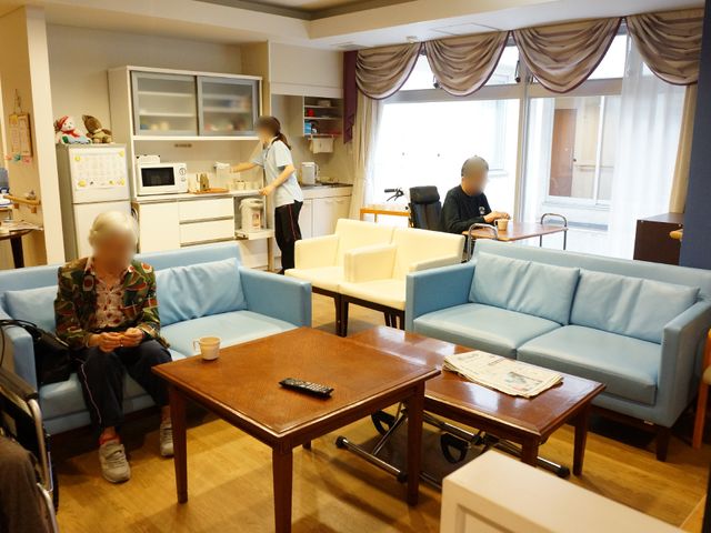 共有リビング空間 ぬくもり感漂う空間 居心地の良い共有リビングで、利用者がリラックスしながら過ごしている様子が見受けられます。スタッフも優しく対応しており、温かい雰囲気が漂っています。