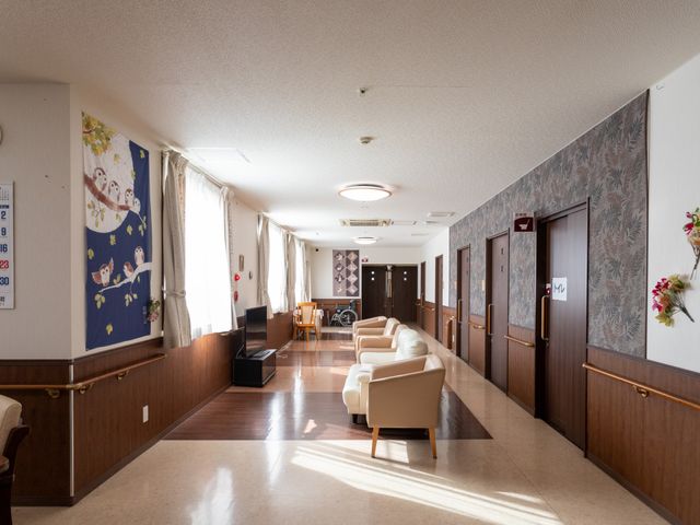明るい廊下の様子 廊下 広々とした廊下は明るく、手すりが設置されており、壁には絵が飾られています。ソファには何名かの方が座っておられ、安らぎのひとときを過ごされている様子です。