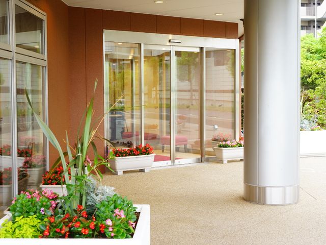 明るいエントランスの風景 施設入り口 花々が彩るエントランスは、訪れる方々を暖かくお迎えします。ゆとりのスペースが心地よい出会いの場を提供しています。