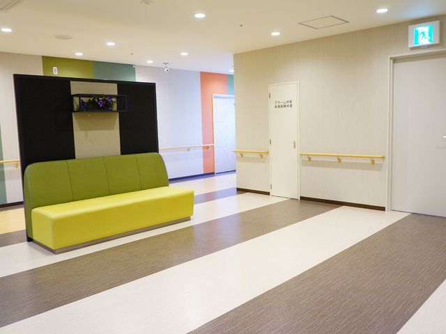 カラフルな廊下とソファ エレベーターホール 落ち着いた色合いのロビーには、鮮やかなソファが置かれ、モダンな雰囲気を醸し出しています。壁には手すりが設けられ、利用者の移動をサポートしています。